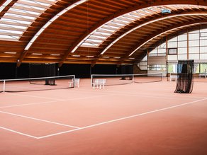 Overzicht van de indoor tennisbanen bij Dekker Warmond, met rode tennisvloeren, een houten gewelfd dak en veel natuurlijk daglicht.