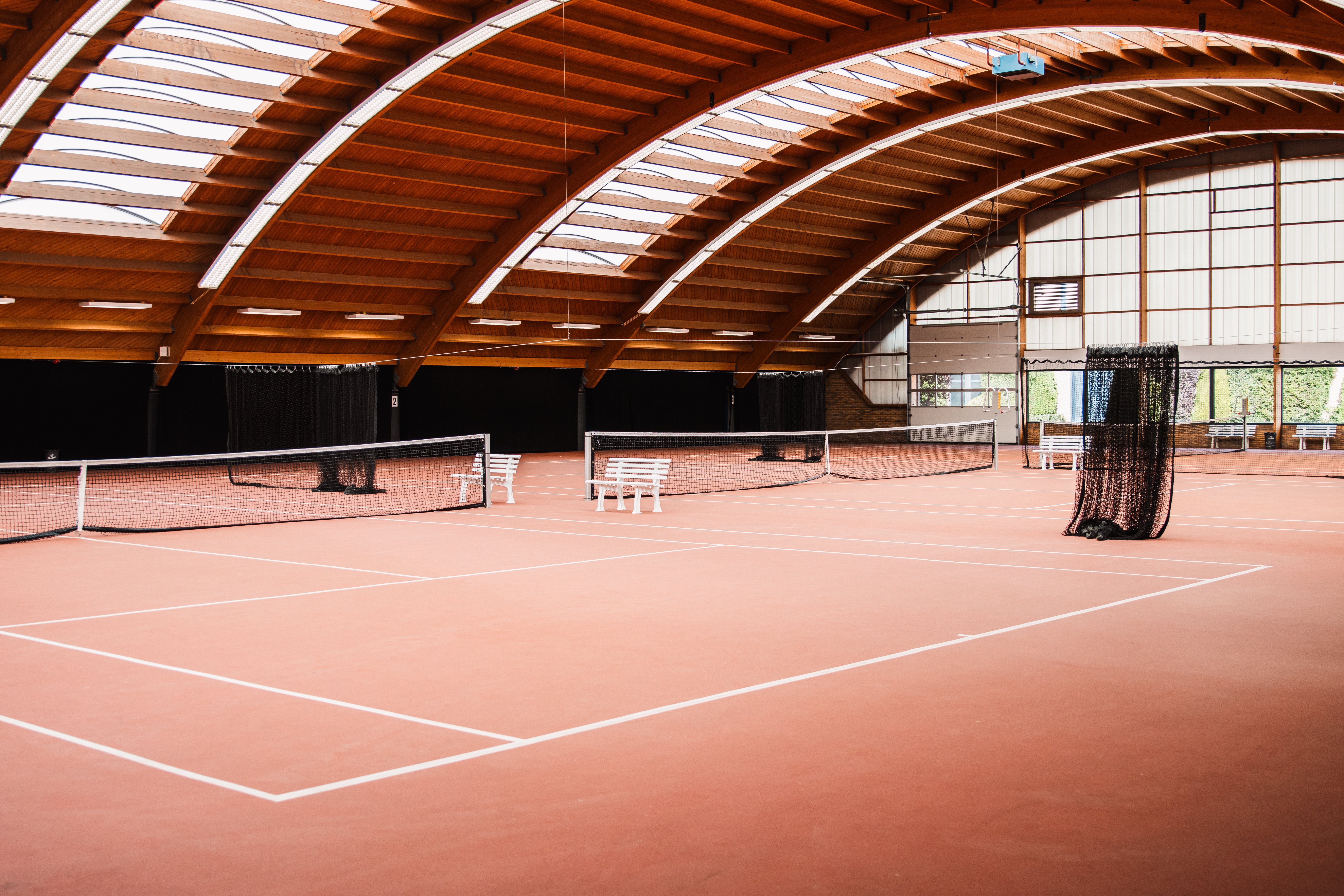 Overzicht van de indoor tennisbanen bij Dekker Warmond, met rode tennisvloeren, een houten gewelfd dak en veel natuurlijk daglicht.