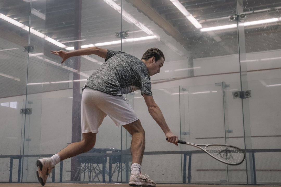 Squashspeler in actie op een indoor squashbaan bij Dekker Warmond, terwijl hij met zijn racket een lage bal probeert te slaan.