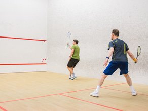 Twee mannen spelen squash op een indoor squashbaan bij Dekker Warmond. Met hun rackets in de hand zijn ze volop in actie tijdens een sportieve wedstrijd.