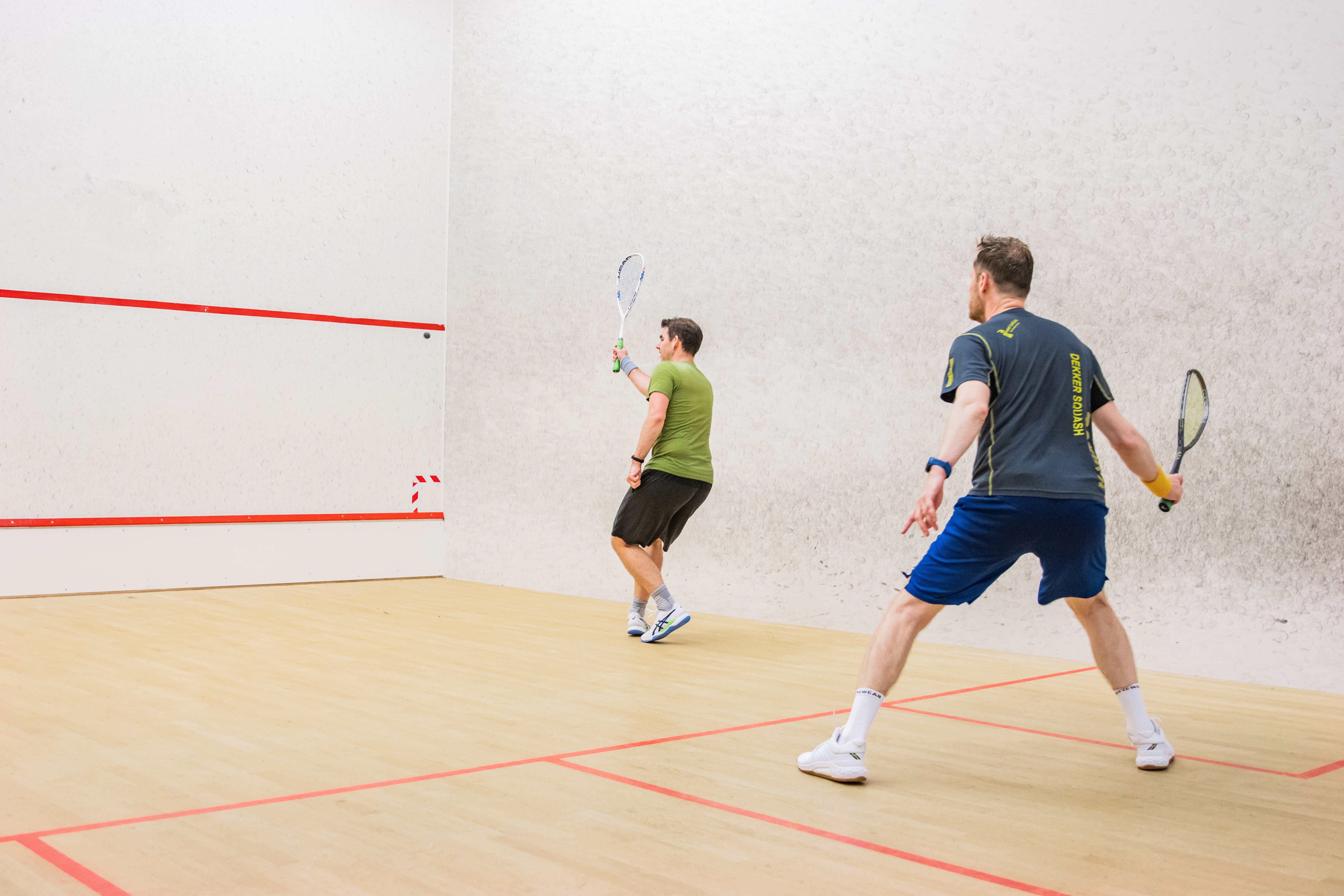 Twee mannen spelen squash op een indoor squashbaan bij Dekker Warmond. Met hun rackets in de hand zijn ze volop in actie tijdens een sportieve wedstrijd.