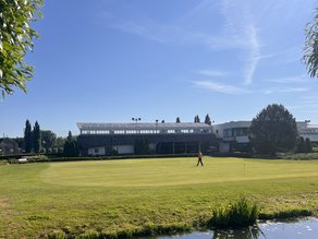Zonnig uitzicht op de golf putting green bij Dekker Warmond, met een vijver op de voorgrond en het sportcomplex op de achtergrond.