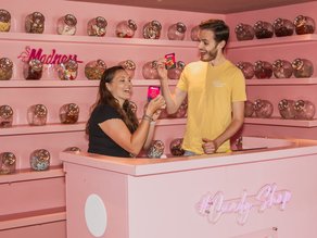 Een lachende man en vrouw genieten van snoepjes in de roze Candy Shop van Madness bij Dekker Zoetermeer, omringd door glazen potten vol kleurrijk snoep.
