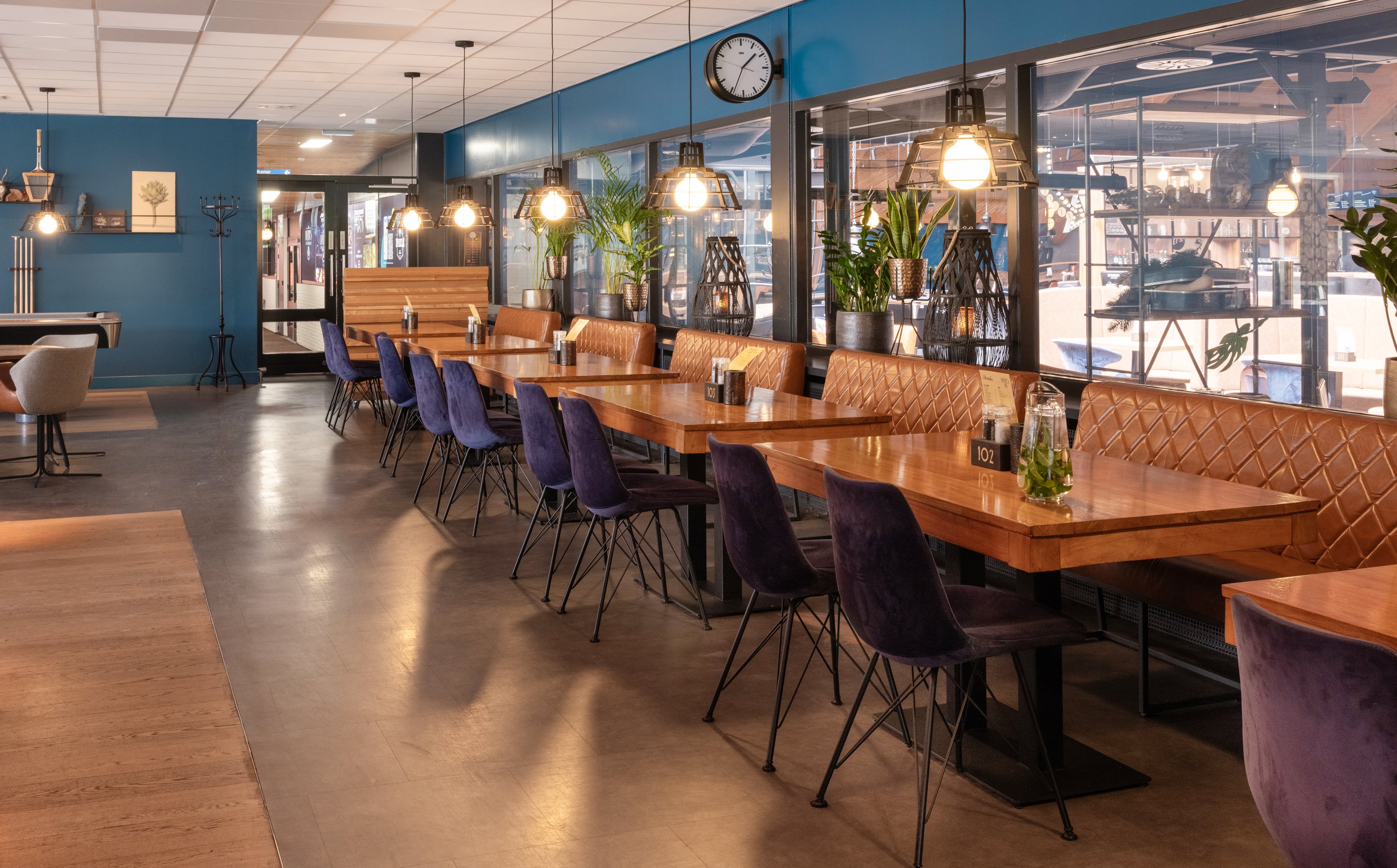 Sfeervol interieur van de sportbar bij Dekker Warmond met houten tafels, comfortabele zitbanken, paarse stoelen en uitzicht op de sportfaciliteiten.