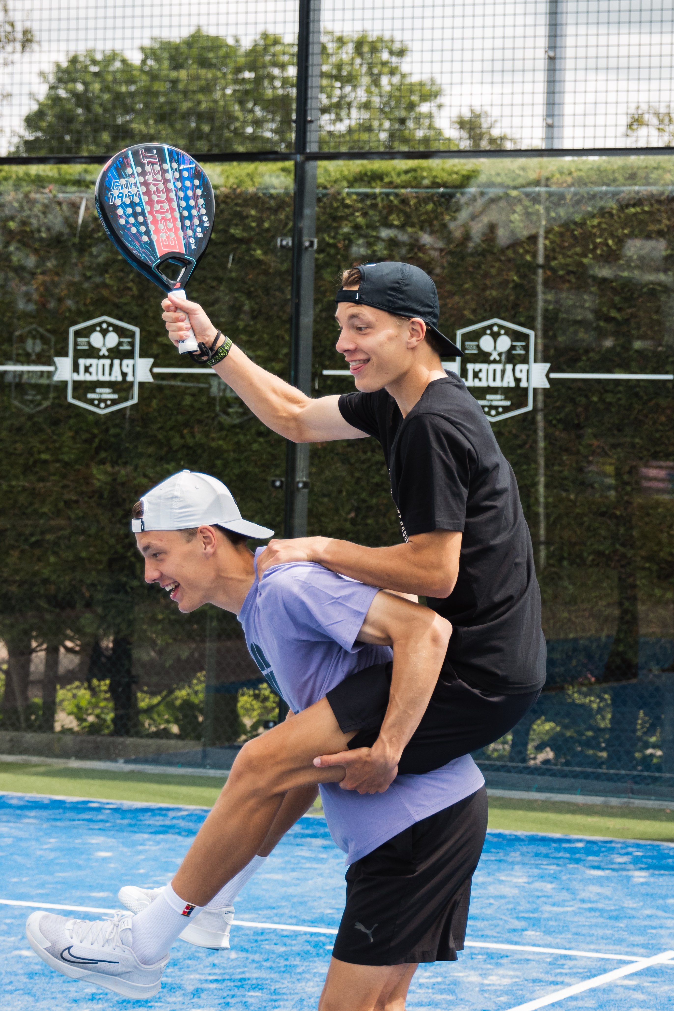 Twee vrolijke jongens maken plezier op de blauwe padelbaan bij Dekker Warmond. Eén jongen zit op de rug van de ander en houdt lachend een padelracket omhoog.
