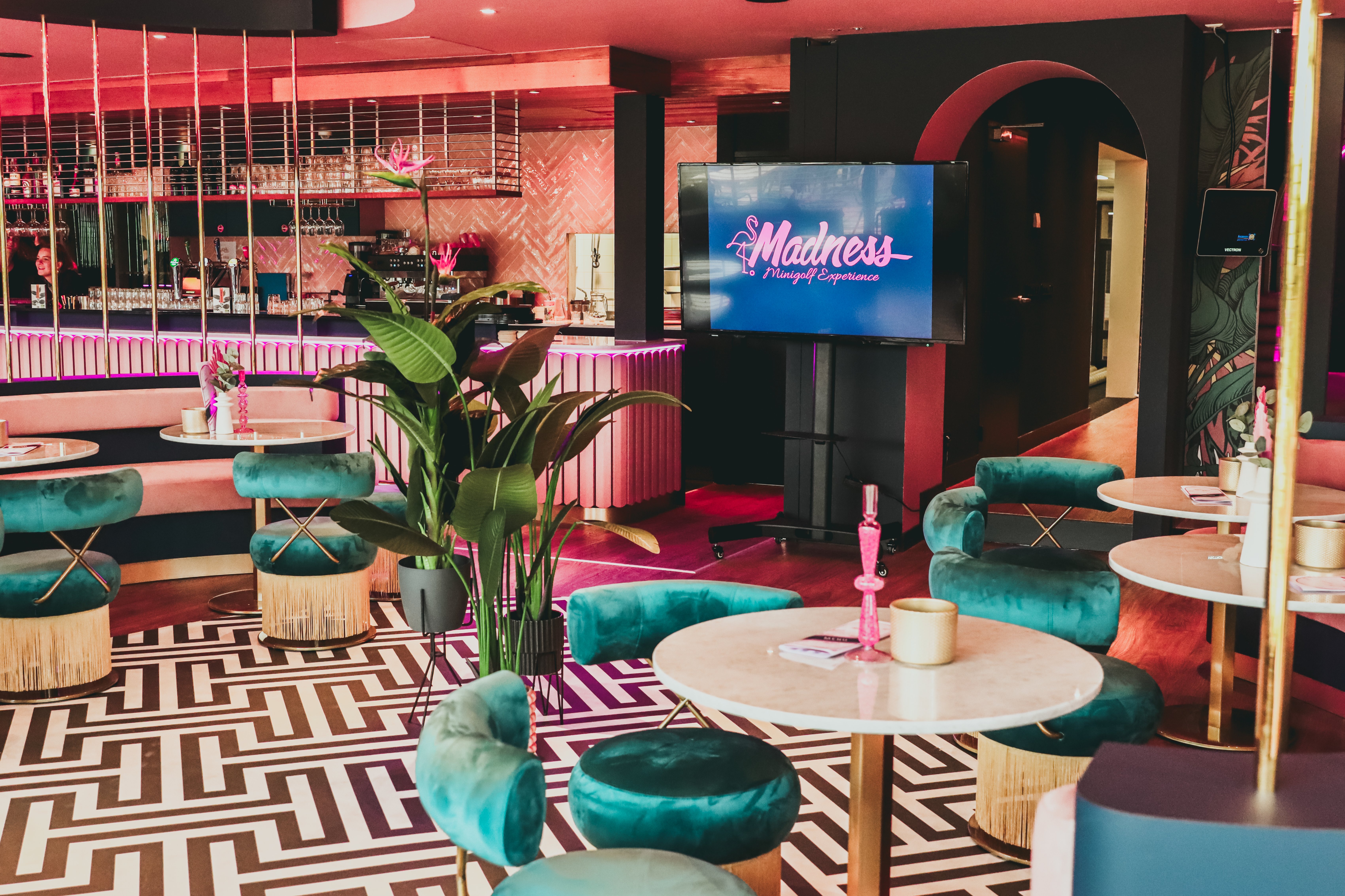 Sfeervolle Madness Minigolf Lounge bij Dekker Warmond, met roze verlichting, turquoise velvet stoelen, ronde tafels en een kleurrijk interieur.