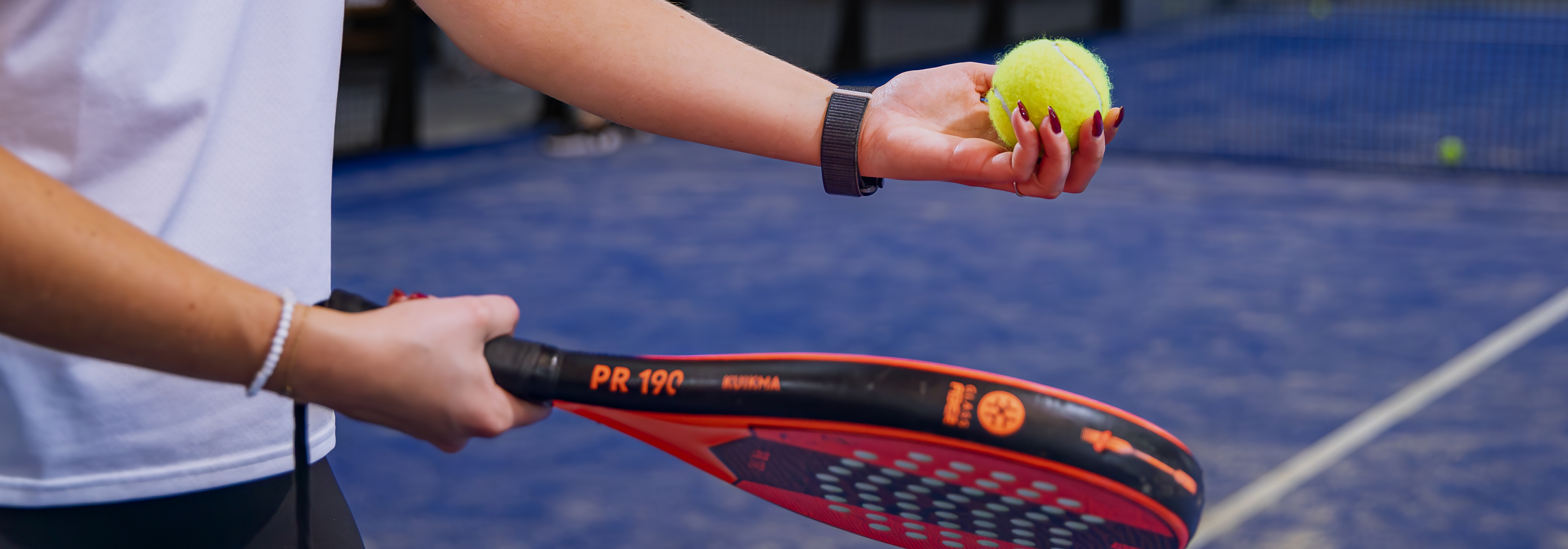 Close-up van een padelspeelster met een padelracket en een gele padelbal op de blauwe padelbaan bij Dekker Warmond.