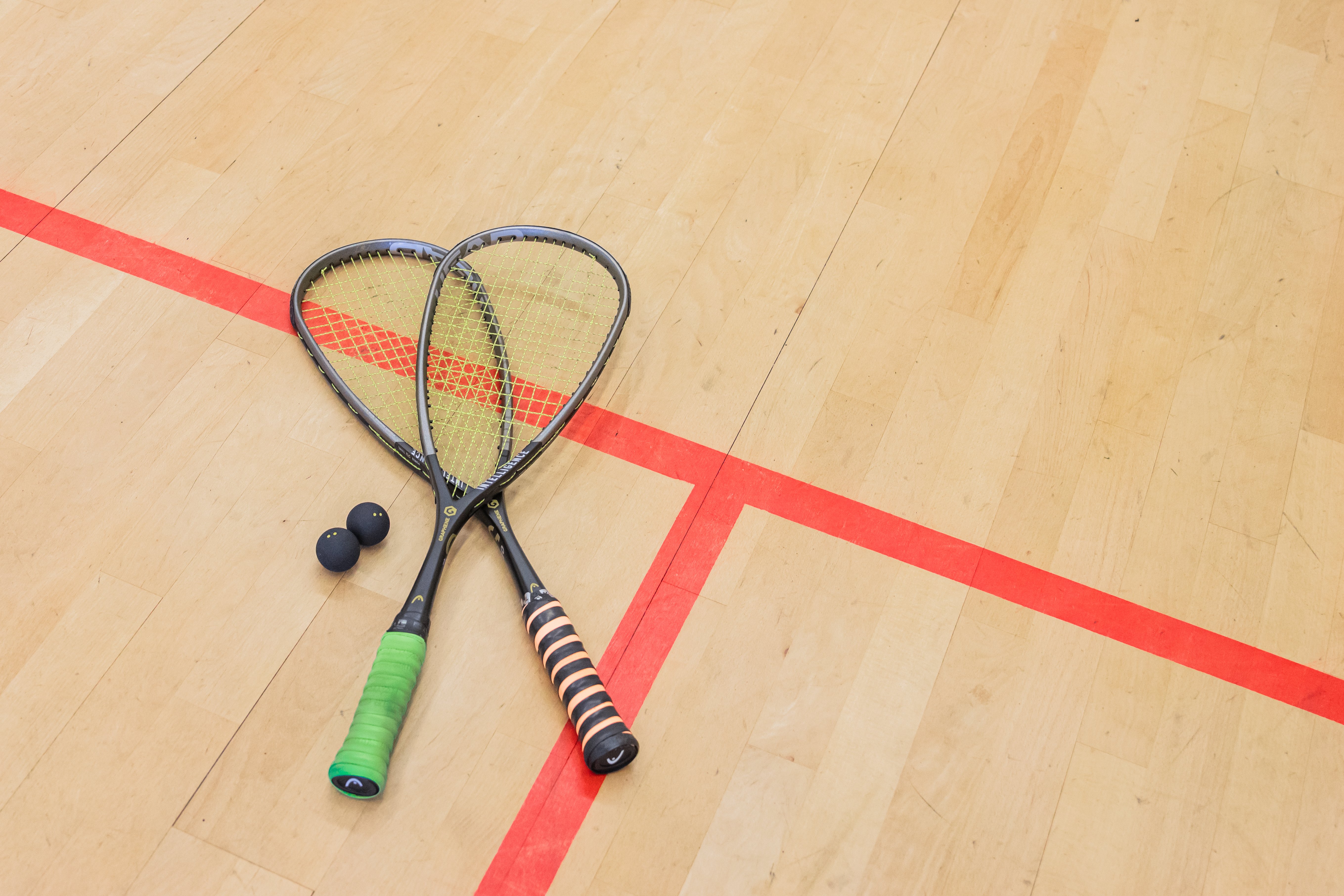 Twee gekruiste squashrackets en twee zwarte squashballen op de houten squashbaan bij Dekker Warmond. De rode belijning van de baan is duidelijk zichtbaar.