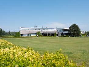 Uitzicht op de groene putting green en het clubgebouw van Dekker Warmond, waar golfers oefenen op een zonnige dag.