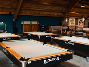 Sfeervolle poolruimte bij Dekker Warmond met meerdere professionele pooltafels, houten plafondbalken, warme verlichting en een gezellige zithoek bij Lucky’s Bowling.