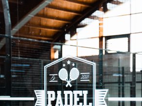 Padel-logo van Dekker Warmond op de glazen wand van een indoor padelbaan, met op de achtergrond de blauwe padelbanen.