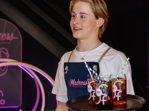 Gastvrije medewerker van Madness Minigolf Experience serveert een dienblad met kleurrijke drankjes in de sfeervolle, roze verlichte minigolflocatie.