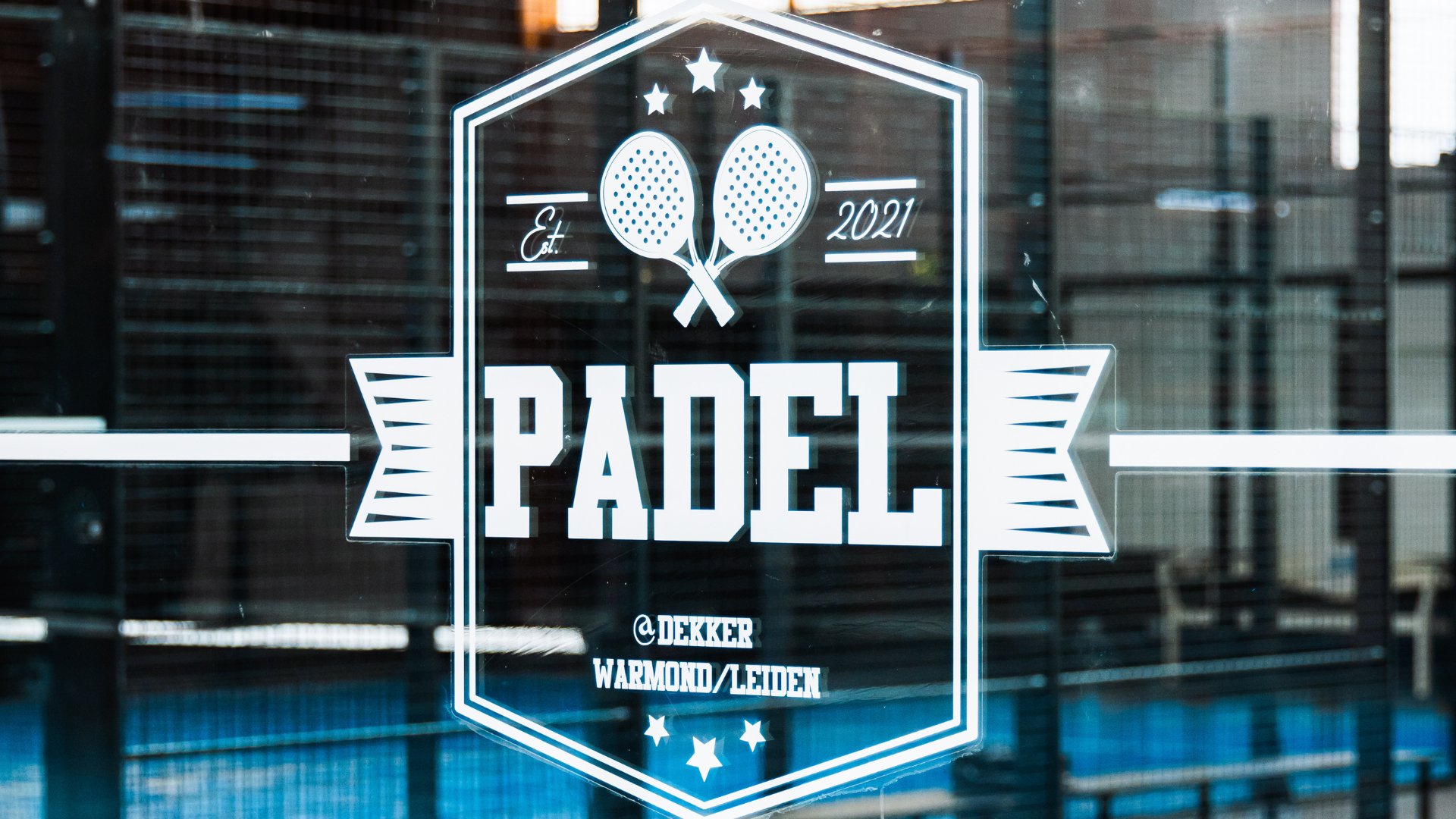 Logo van Padel @Dekker Warmond/Leiden op de glazen wand van een indoor padelbaan bij Dekker Warmond.