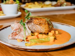 Een mooi opgemaakt vleesgerecht met romige oranje saus, verse groenten, rauwe ham en groene kruiden, geserveerd op een bord bij Dekker Warmond.