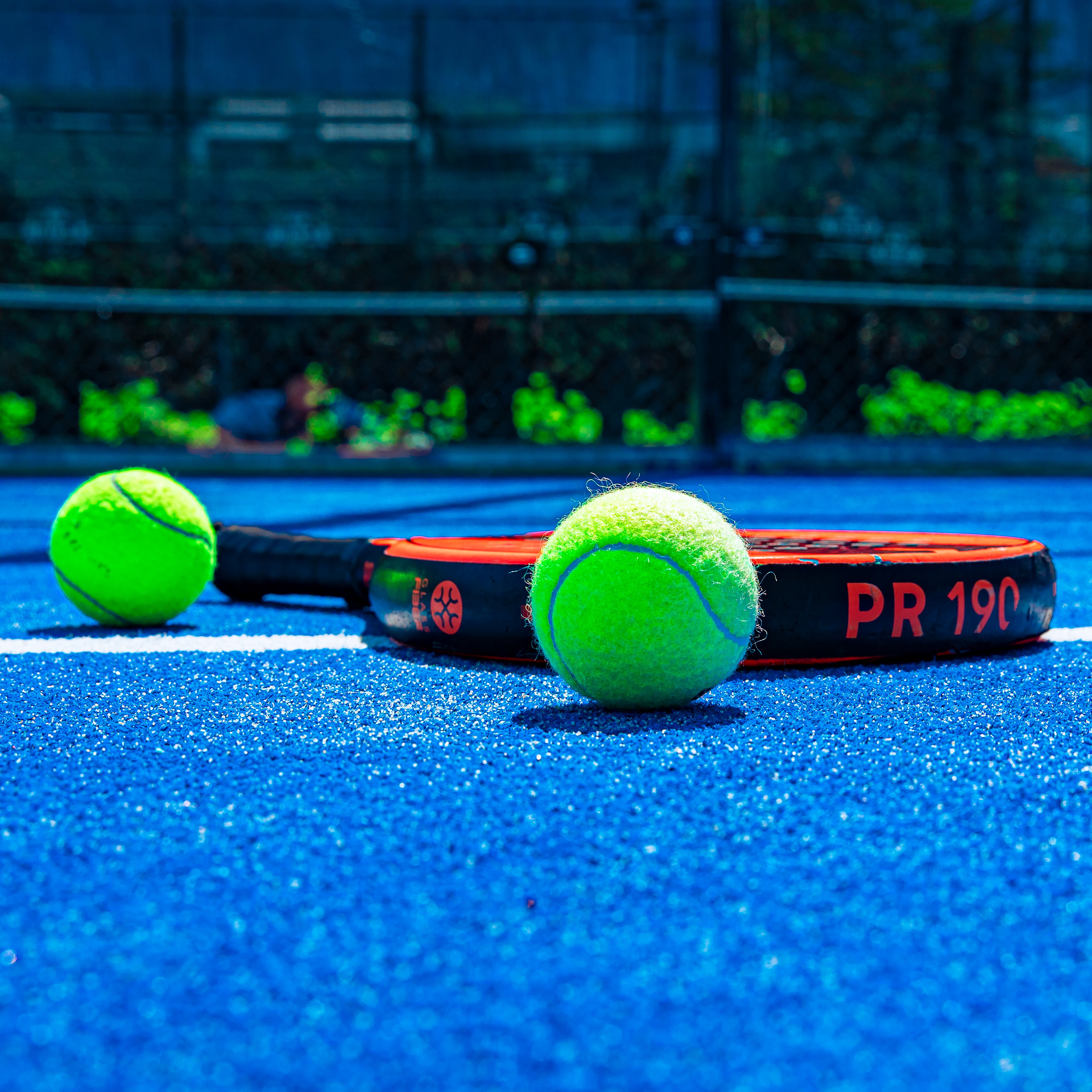 Close-up van een zwart-rood padelracket en twee felgele padelballen op de blauwe padelbaan bij Dekker Warmond, met het hekwerk op de achtergrond.