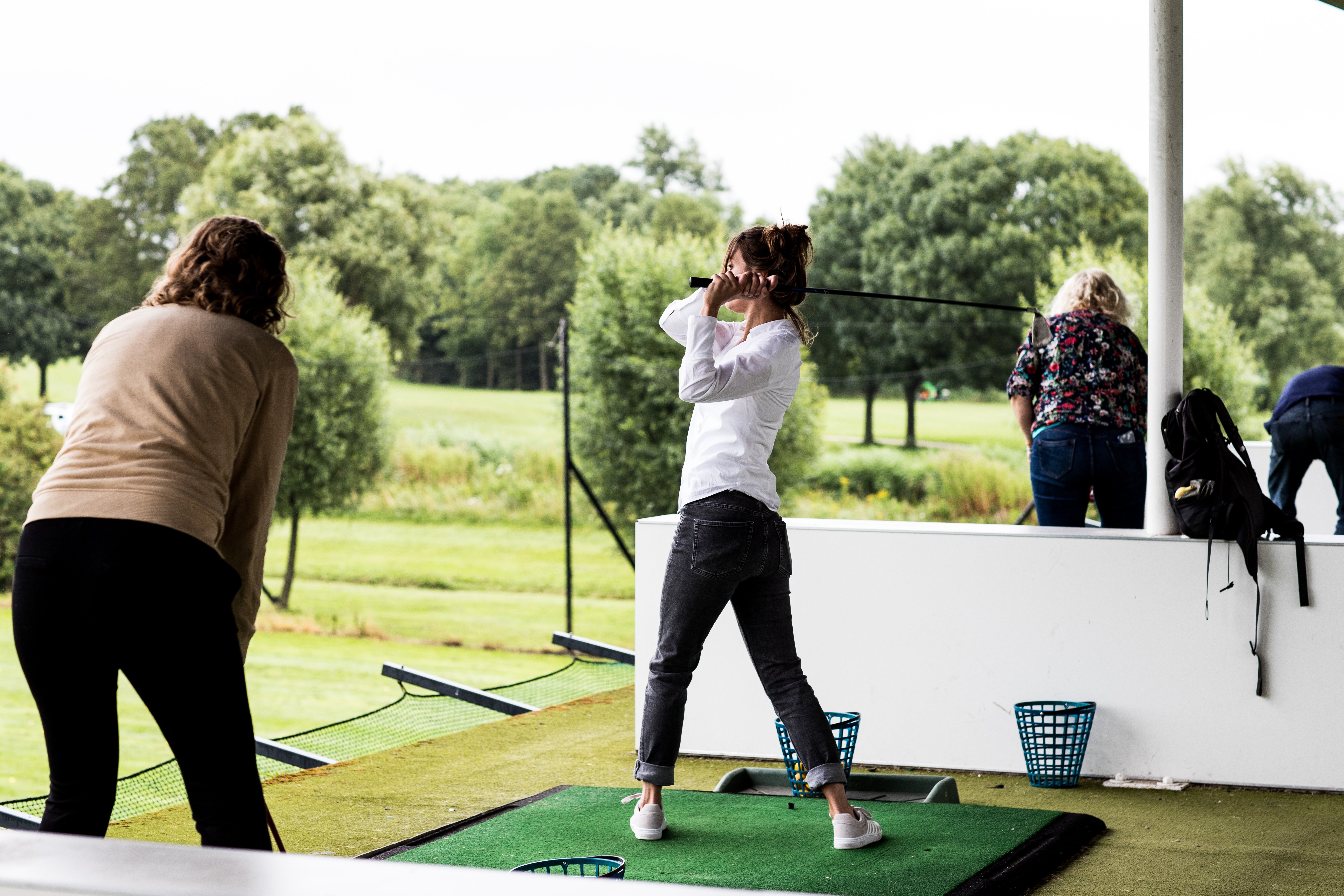 Dames oefenen hun golfswing op de driving range bij Dekker Warmond, met uitzicht op de groene golfbaan.