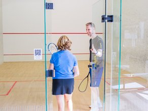 Een man en een vrouw staan met squashrackets op de indoor squashbaan bij Dekker Warmond. Ze praten en lachen samen na het squashen.