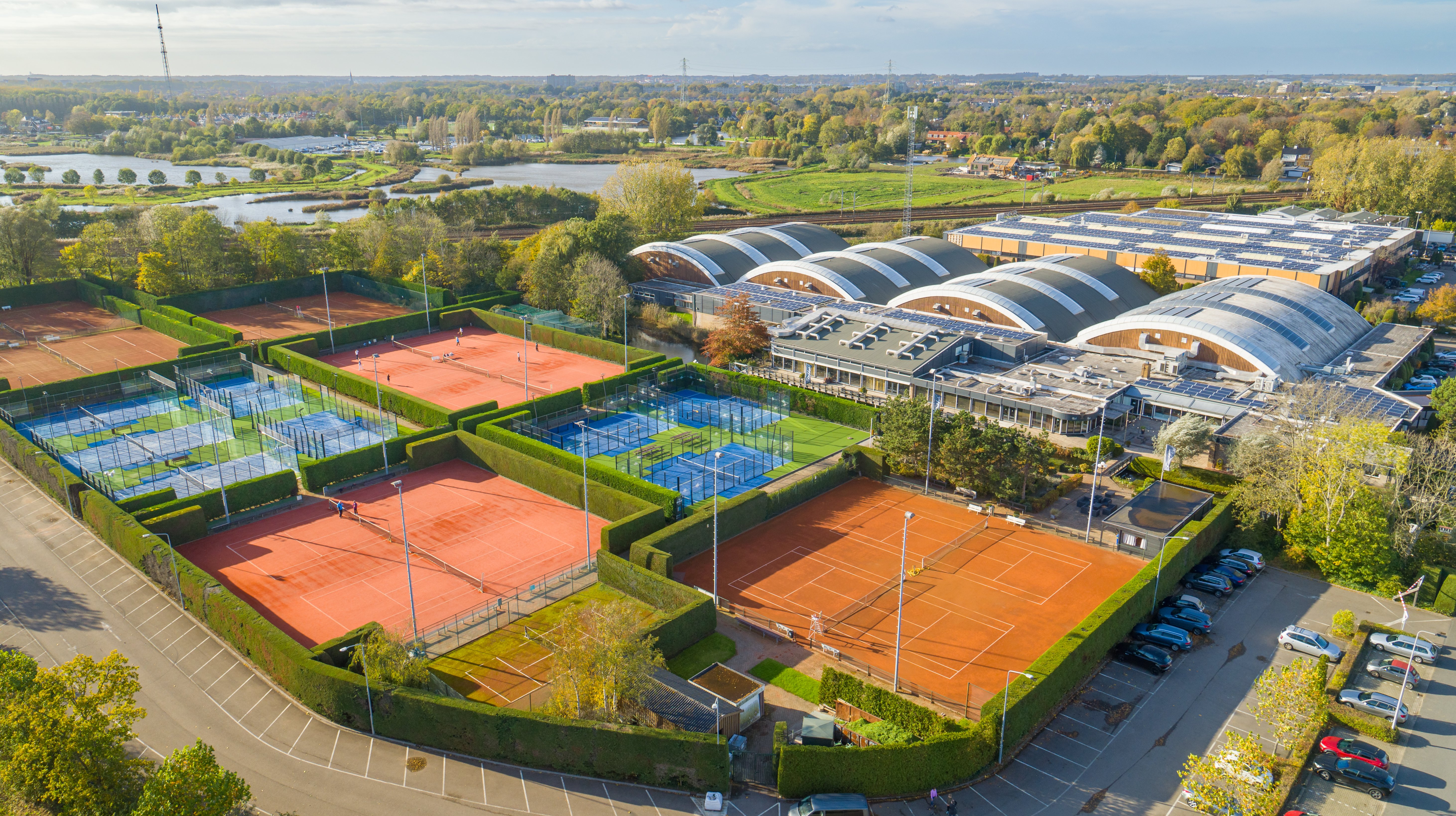 Luchtfoto van Dekker Warmond met buitenbanen voor tennis en padel, de overdekte sporthallen, parkeergelegenheid en een groene omgeving met waterpartijen.