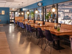 Sfeervolle horecaruimte bij Dekker Warmond met houten tafels, comfortabele paarse stoelen, cognackleurige zitbanken, warme hanglampen en grote ramen met uitzicht op de aangrenzende ruimte.