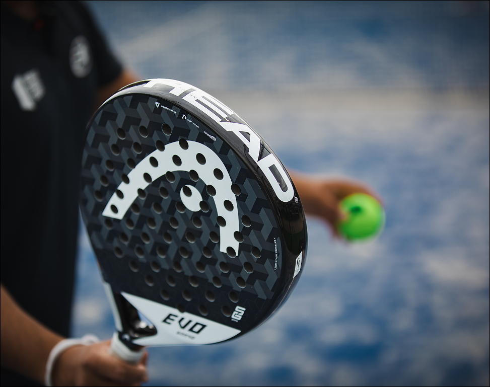 Close-up van een HEAD padelracket met een groene padelbal op een blauwe padelbaan bij Dekker Warmond.