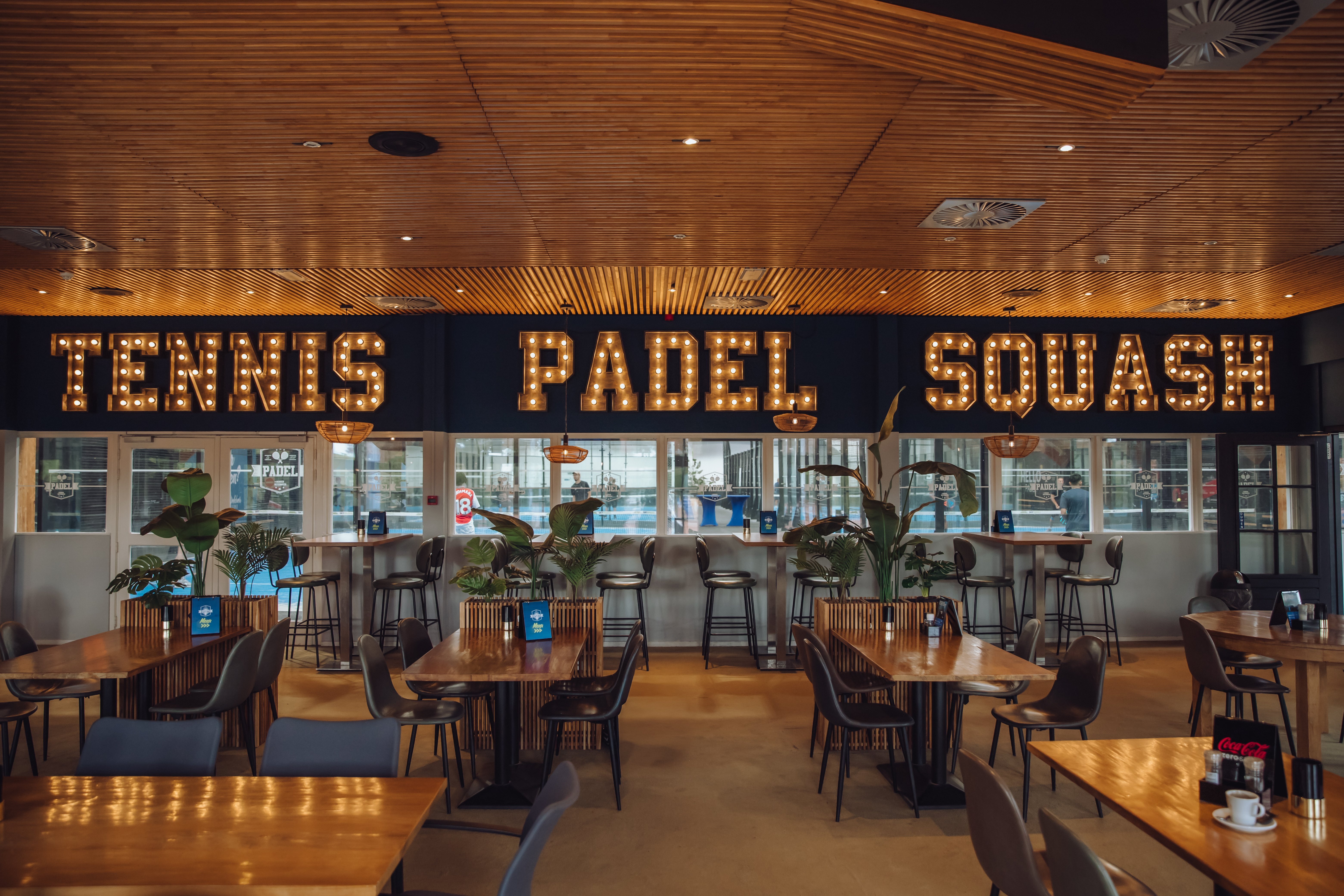 Sfeervolle sportbar bij Dekker Warmond met houten tafels, comfortabele zitplaatsen en verlichte letters Tennis, Padel en Squash, met uitzicht op de indoor padelbanen.