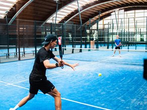 Twee sporters spelen padel op een blauwe indoor padelbaan bij Dekker Warmond. Een speler in zwarte sportkleding staat klaar om de bal terug te slaan.